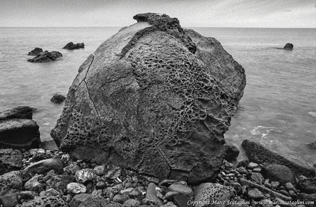 Monochrome view of a large rock on the beach with the ocean in the background under natural lighting.