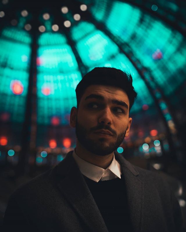 A stylish man with a beard poses indoors, with blurred lights creating a moody atmosphere.
