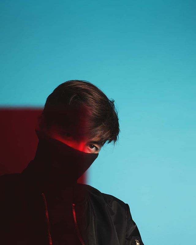 A man in a black jacket and face covering stands against a blue and red backdrop in a dramatic editorial style image.