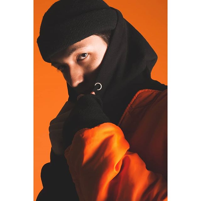A man in a black hoodie and beanie poses against an orange background with a serious expression.