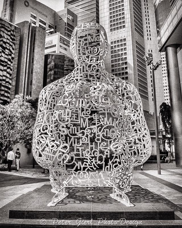 A sculpture composed of letters stands tall against a backdrop of skyscrapers in an urban setting.