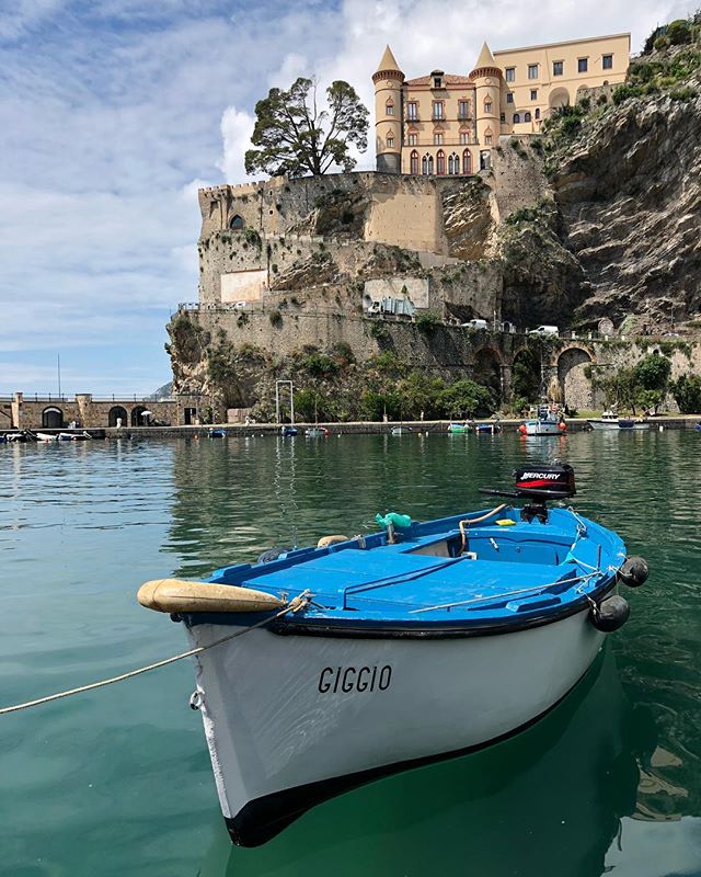 A blue and white boat is moored in the calm waters of an Italian harbor with a cliffside castle in the background.