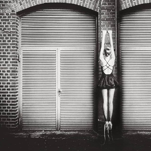 A ballerina in a dark dress and ballet shoes stands with arms raised in front of a brick wall and metal doors.