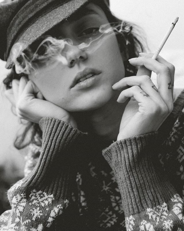 A woman in a hat and sweater is smoking a cigarette in a black and white portrait.