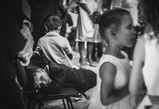 A black and white image captures children at an event, one boy resting on a chair looking at the camera.