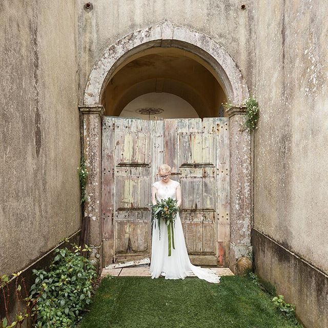 A bride stands with a bouquet in front of an old archway and weathered doors in a serene garden setting.