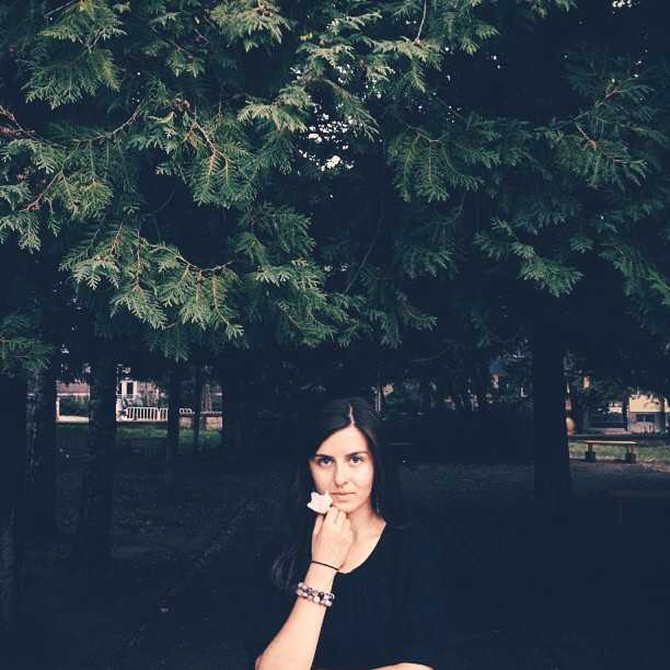 A woman stands peacefully under a tree in a park, holding a white flower, enjoying a moment of serenity.