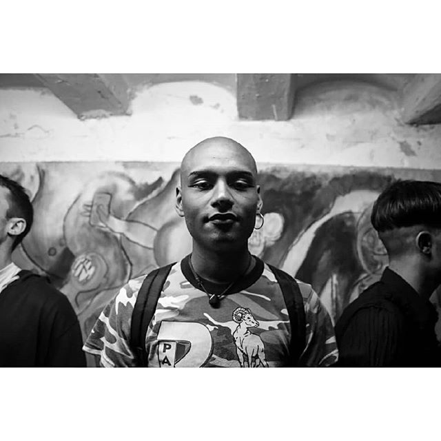 Black and white portrait of a bald man with an earring wearing a patterned t-shirt.