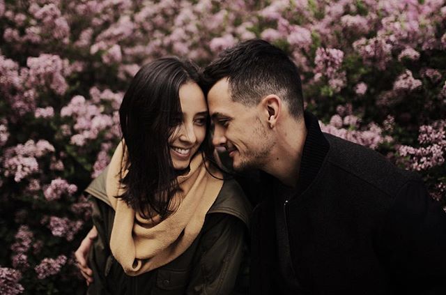 A smiling couple embraces in front of blooming lilac bushes in a romantic outdoor setting.