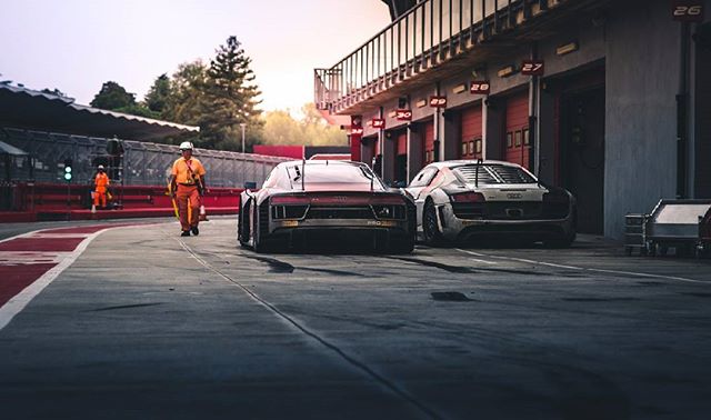 Two Audi R8 race cars sit in the pit lane as a track worker walks by a garage at a racetrack.
