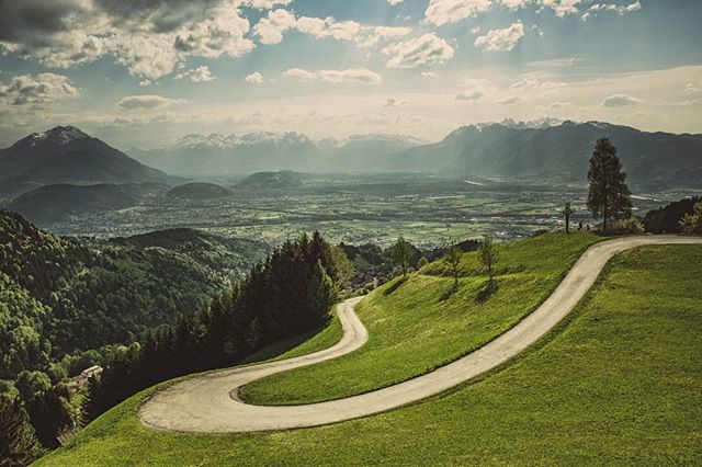 A scenic mountain road winds through lush green hills with snow-capped peaks in the distance under a partly cloudy sky.