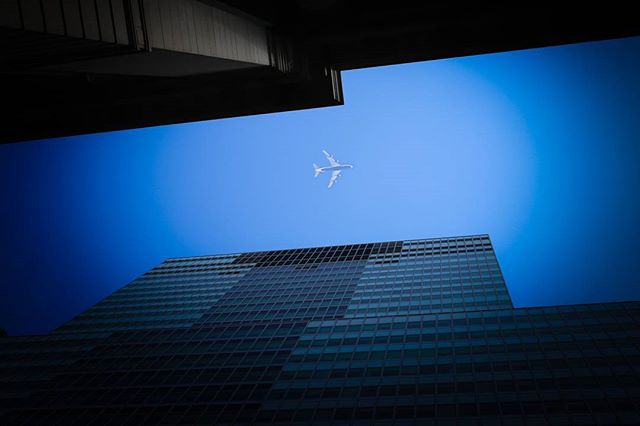 An airplane soars in the blue sky above a modern building, framed by architectural elements in an urban setting.