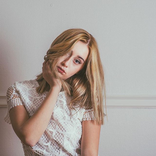 A woman with blonde hair in a lace top poses pensively against a white background.