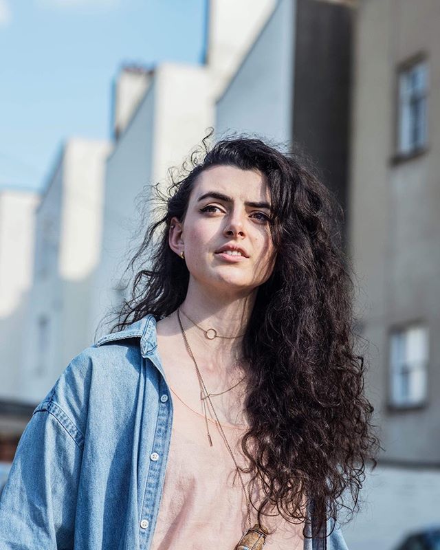 A confident woman in a denim jacket stands in an urban setting, exuding a natural and relaxed vibe.