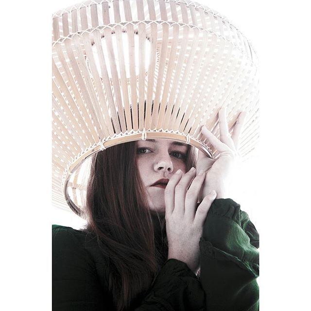 A woman poses with her hands near her face, under a woven lamp shade in a close up indoor shot.