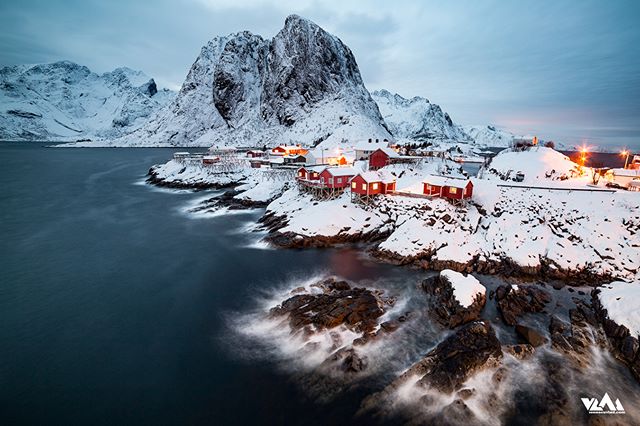 Winter landscape of a village on the coast of Norway with snow-covered mountains in the background.