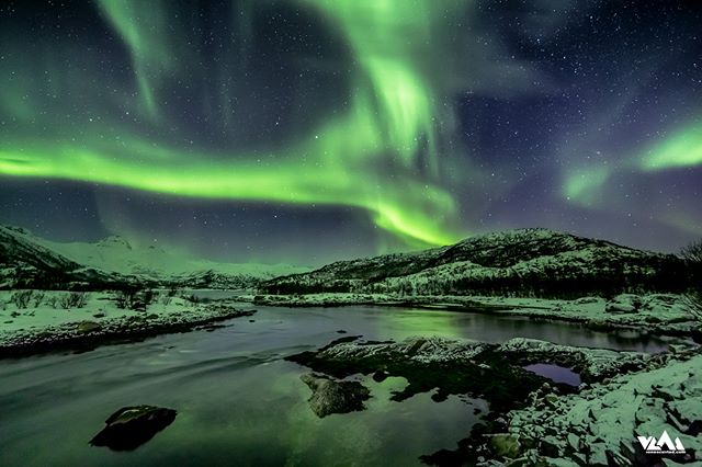 A stunning aurora borealis dances over a serene river in a snowy, mountainous landscape at night.