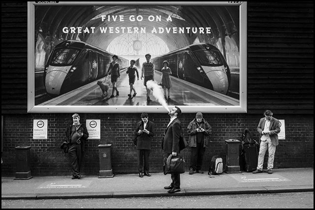 Black and white image of people waiting near a train station billboard, one man is smoking, in an urban scene.