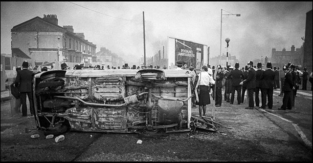A crowd of people including police officers are gathered around an overturned car in a black and white, urban street scene.