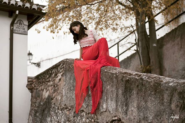 A woman in a red dress sits contemplatively on a stone wall in a European city.