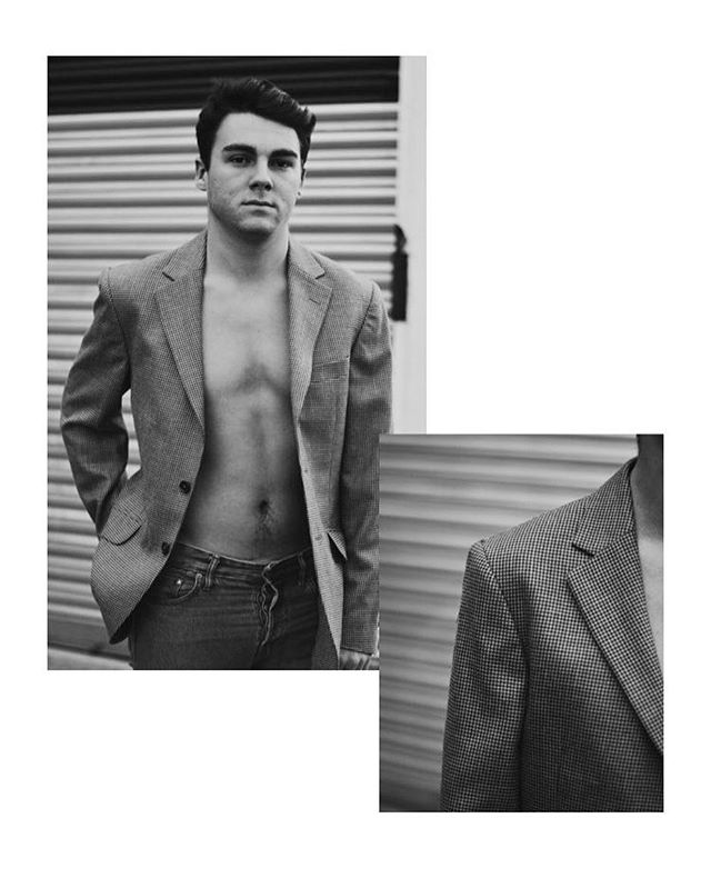Monochrome portrait of a shirtless man wearing a blazer and jeans in an urban setting, styled for editorial use.