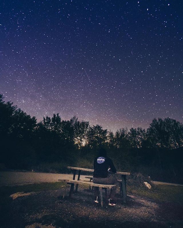 A person wearing a NASA sweatshirt sits on a bench under a starry night sky.