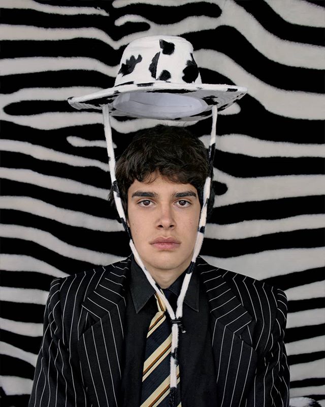 A man wearing a cow print hat and pinstripe suit poses against a zebra print backdrop.