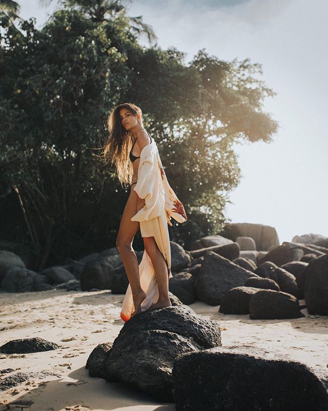 A woman in a bikini and robe stands on a rock at the beach, enjoying the sun and serene atmosphere.