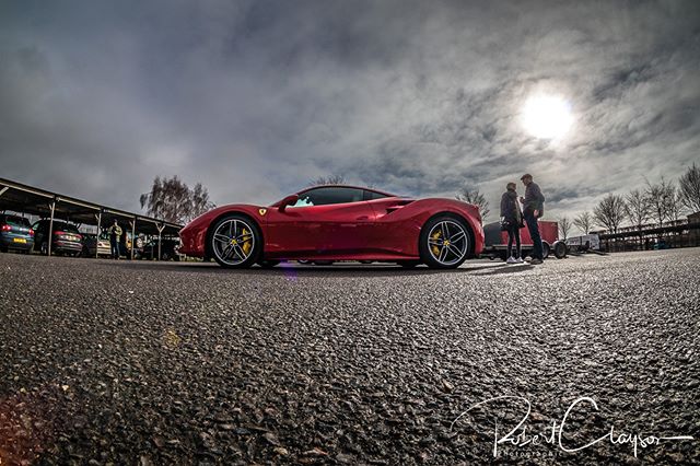 A red Ferrari stands out in a parking lot, with a couple nearby under a cloudy sky.