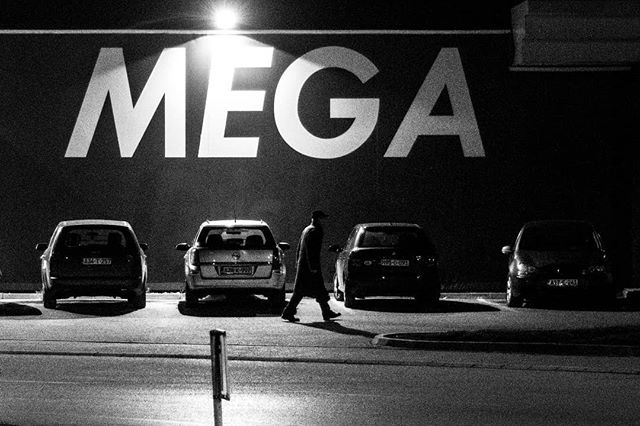 A black and white image of a man walking in front of parked cars and a building with a large MEGA sign.
