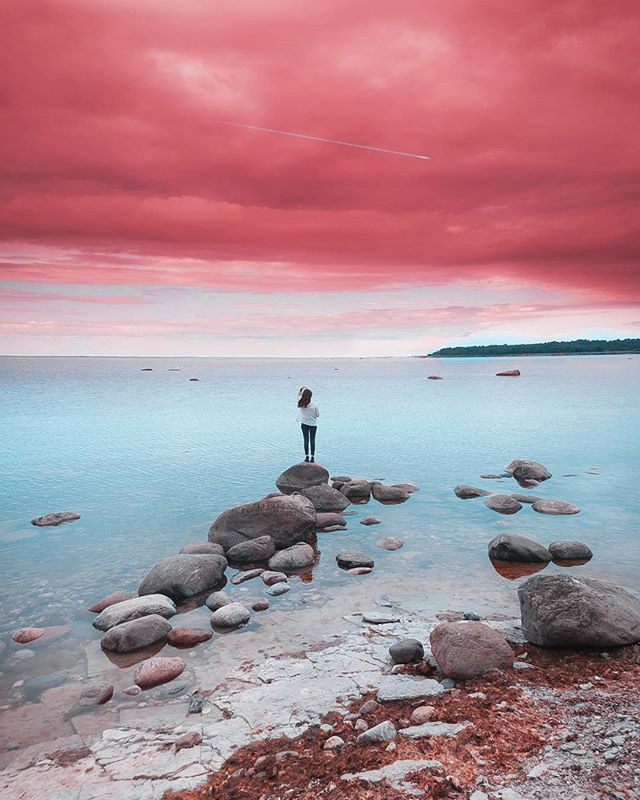 A solitary woman stands on rocks in the ocean against a pink sky, creating a serene and dreamy seascape.