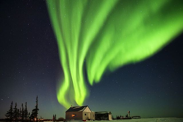 A stunning aurora borealis illuminates the night sky above a remote cabin in a winter wonderland.