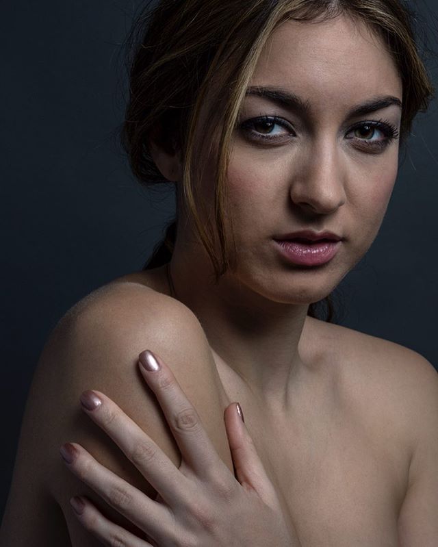 A woman with light skin poses in a soft-lit studio portrait, showing her bare shoulder and hand.
