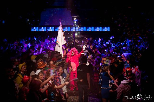 A vibrant crowd celebrates with a person in costume holding a flag at an indoor event.