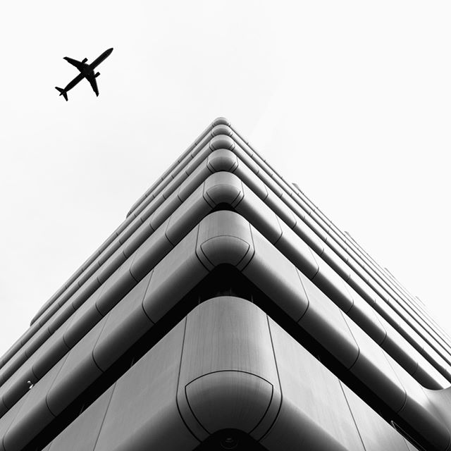 An airplane flies over a modern building, showcasing a blend of urban life and architectural design in grayscale.