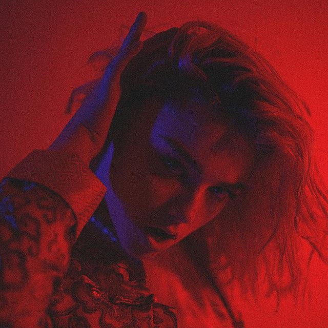 A person with long hair poses in a dramatic red and blue lighting, creating a moody atmosphere.