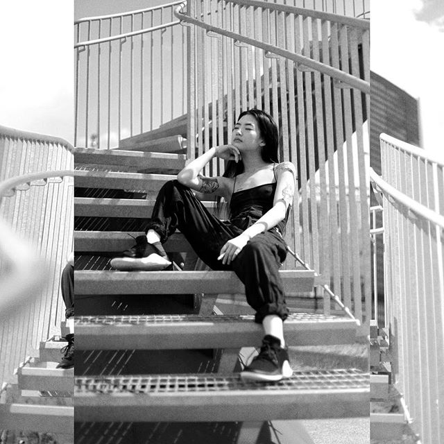 A woman in black sits pensively on metal stairs in a black and white urban setting, bathed in soft light.