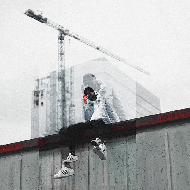 A man sits on a rooftop in an urban setting, wearing sneakers in a moody, monochromatic scene with construction in the background.