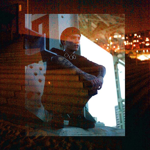 A stylish man with tattoos and a choker sits in an urban setting, creating a moody and mysterious vibe.