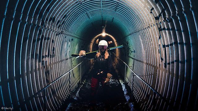 A skier walks through a bright, corrugated metal tunnel carrying skis on his shoulder, creating a moody image.