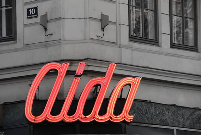 A bright neon “Aida” sign adorns a building in an urban scene, evoking a classic retro vibe.