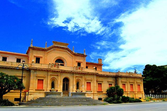 A classical-style building with a grand facade and staircase, set against a bright blue sky with fluffy white clouds.