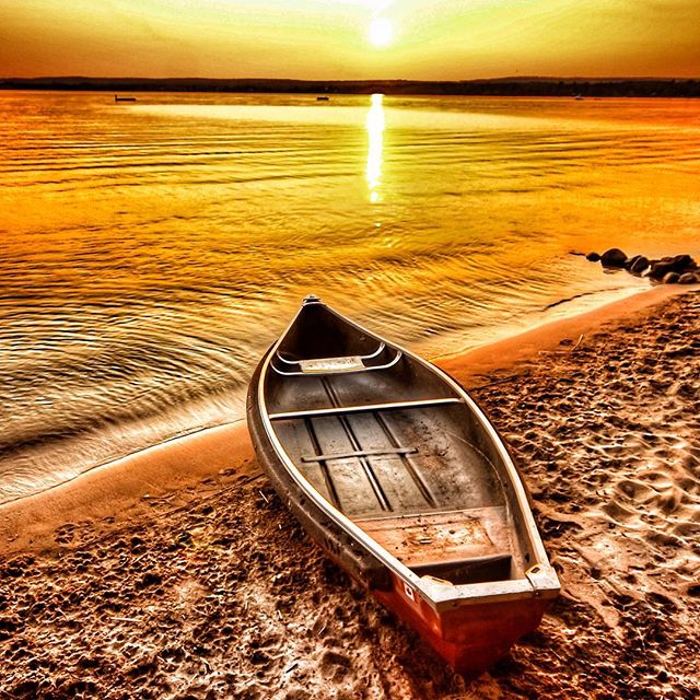 A canoe rests on a sandy beach during a beautiful sunset over a calm lake.