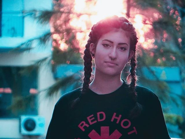 A woman with braids and a nose ring poses outdoors in soft light.