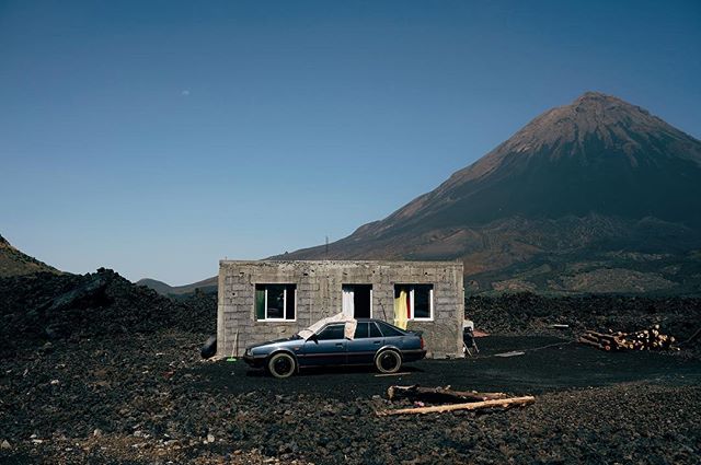 A blue car sits in front of a block building with a volcano looming in the background.