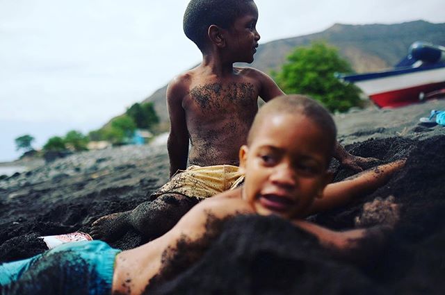 Two children are playing on a black sand beach, covered in sand and enjoying their time.