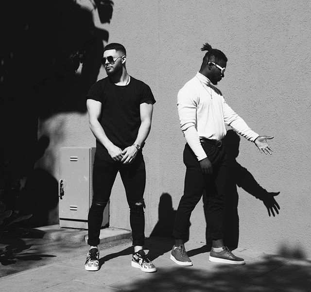 Two stylish men pose in a monochrome street scene with strong shadows, showcasing urban fashion.