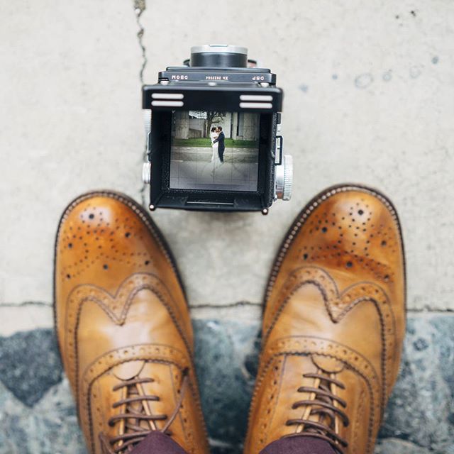 A pair of brown brogue shoes are seen next to a vintage camera that is capturing a loving wedding couple. 