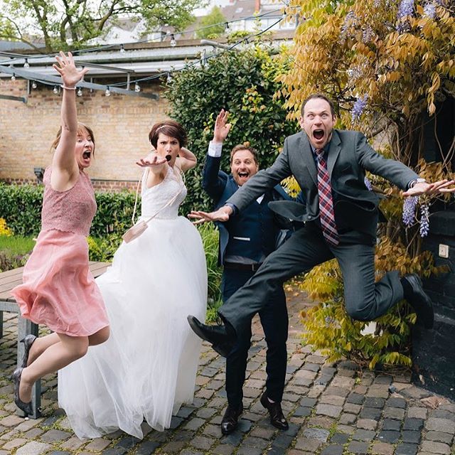 A bride, groom, and two guests jump for joy in a candid outdoor wedding celebration.