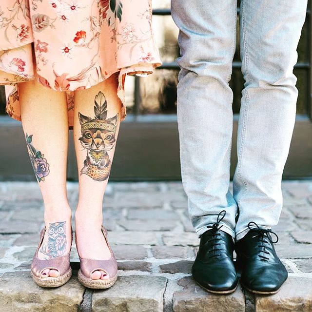 A couple stands together, showcasing unique tattoos and stylish footwear on a brick surface.